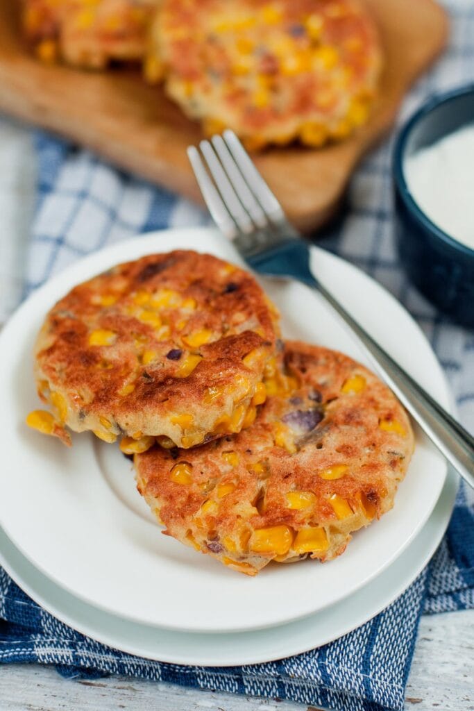 Two sweetcorn fritters and fork are sat on a white plate on top of a blue checked cloth. In the background is a pot of sour cream and two more fritters on a board | Hurry The Food Up