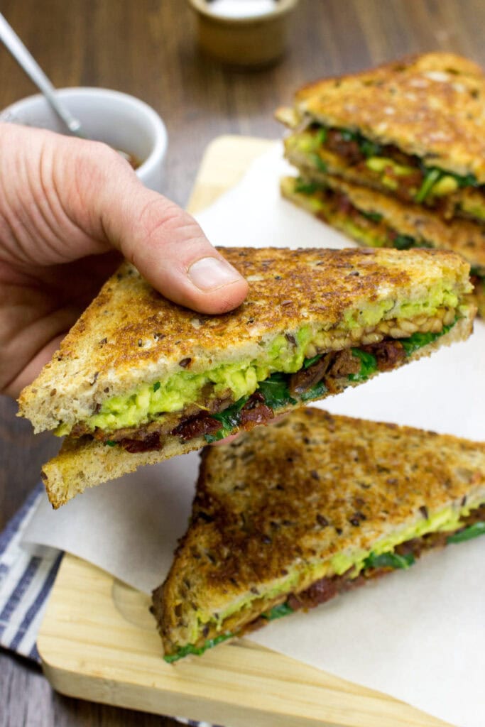 Half of a tempeh sandwich is held up with another below it and two in the distance | Hurry The Food