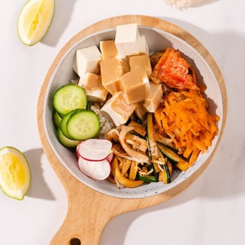 A birds-eye view of a marbled table with a wooden chopping board on top. On the board is a bowl of vegan bibimbap. Next to the bowl are two lemon wedges and a cream cloth | Hurry The Food Up