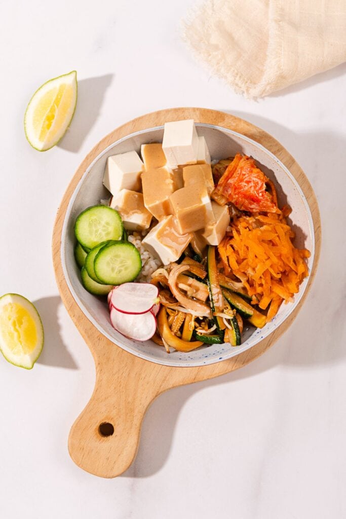 A birds-eye view of a marbled table with a wooden chopping board on top. On the board is a bowl of vegan bibimbap. Next to the bowl are two lemon wedges and a cream cloth | Hurry The Food Up