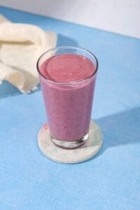 In the centre of a blue table is a white ceramic coaster with a tall glass full of blackberry smoothie. Next to the glass is a linen cloth | Hurry The Food Up