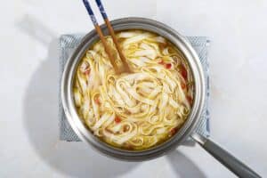 A saucepan of rice noodles in broth, with a pair of chopsticks in it, on a blue and white tea towel. | Hurry The Food Up