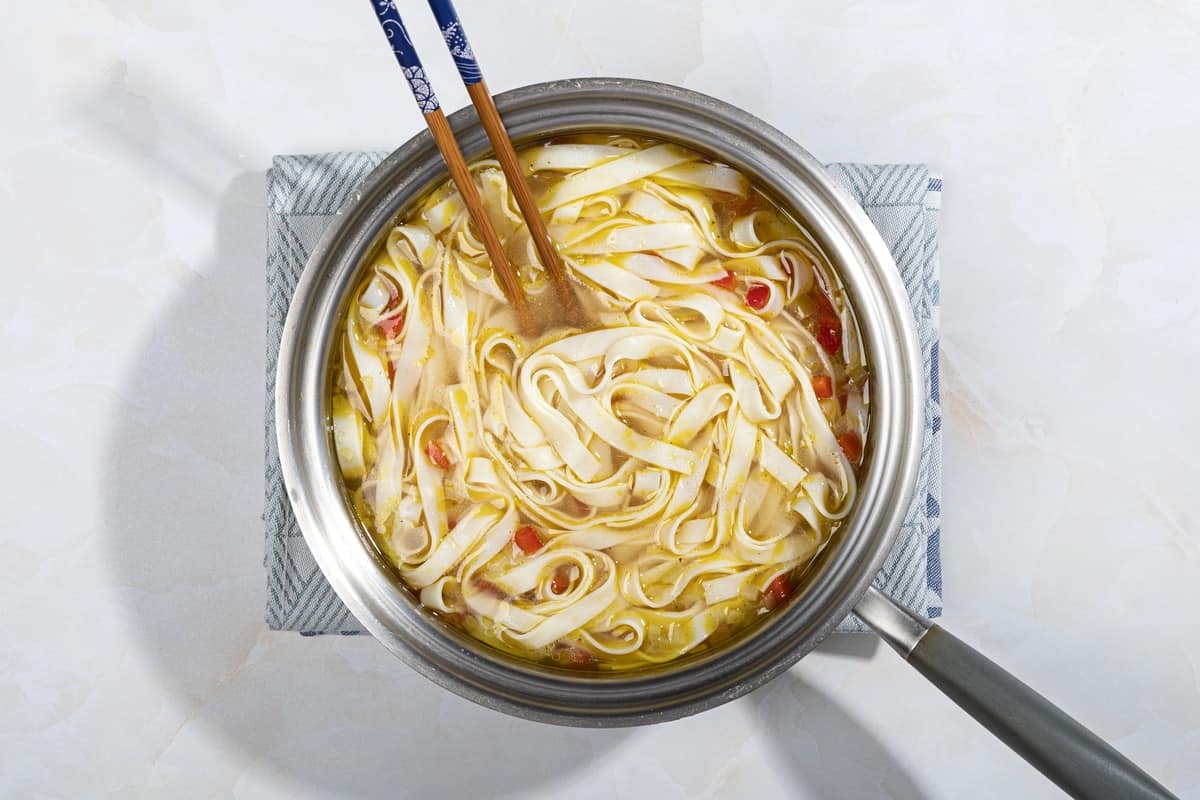 A saucepan of rice noodles in broth, with a pair of chopsticks in it, on a blue and white tea towel | Hurry The Food Up