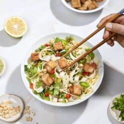 Someone uses chopsticks to pick a piece of tofu out of a bowl of pho, small plates of toppings around. | Hurry The Food Up