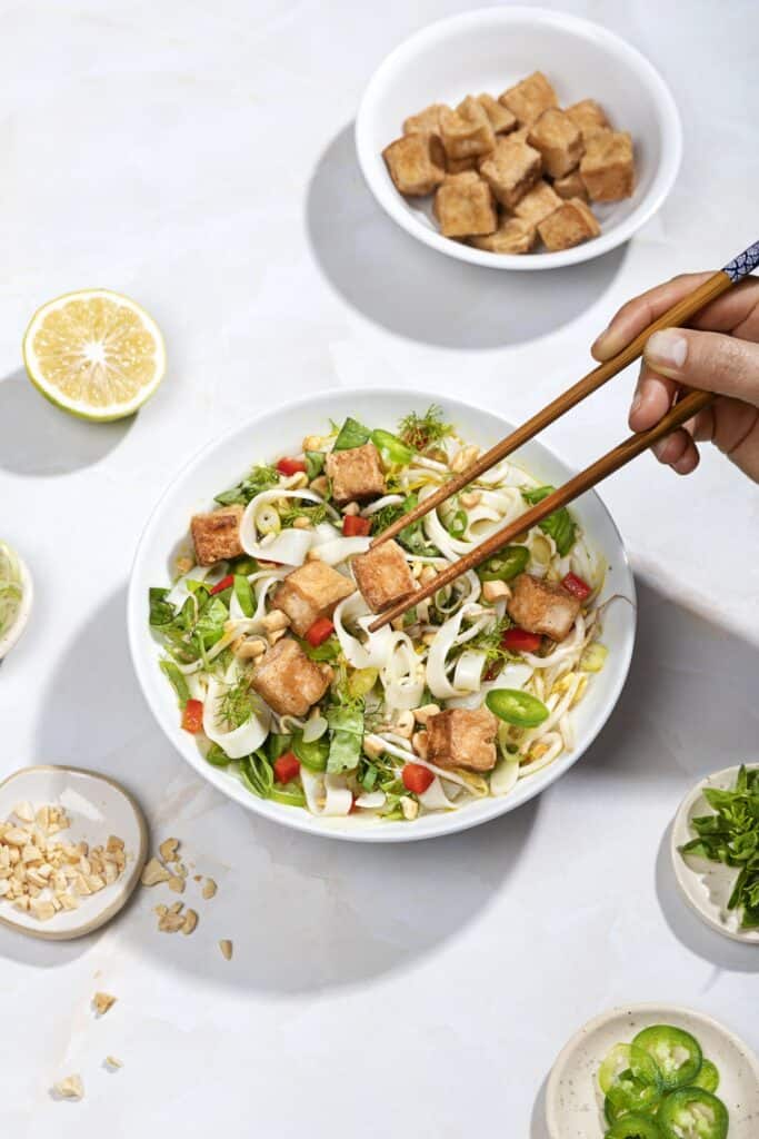 Someone uses chopsticks to pick a piece of tofu out of a bowl of pho, small plates of toppings around. | Hurry The Food Up