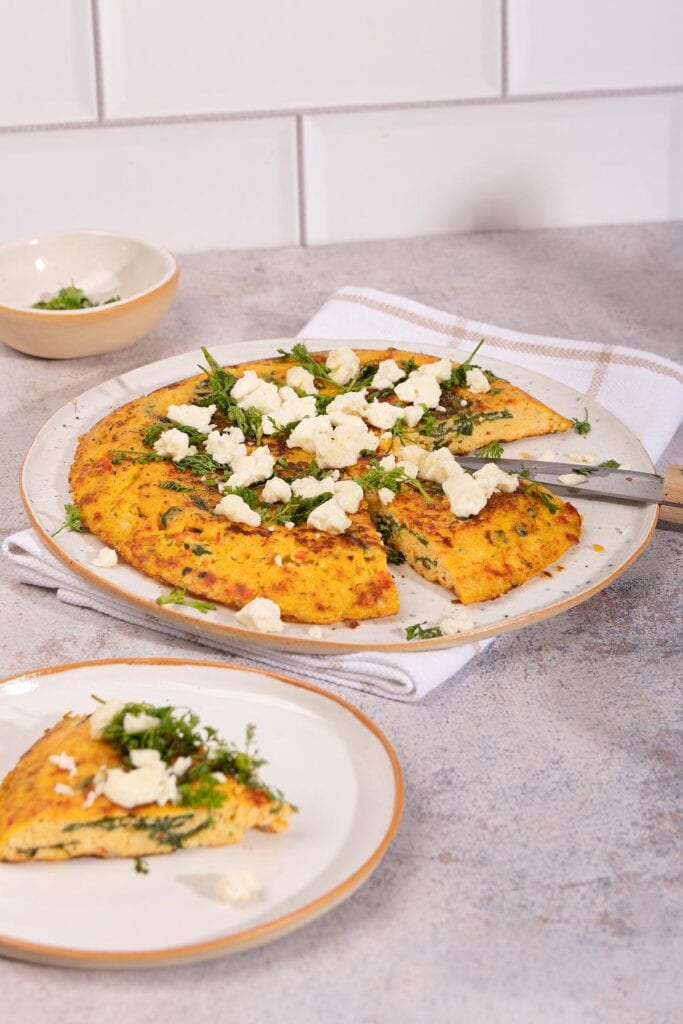 A plate of frittata sits on a white towel. There is a plate with a piece of frittata and a small bowl of herbs on the sides of it | Hurry The Food Up